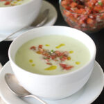 A white bowl of creamy green soup garnished with diced tomatoes, herbs, and a drizzle of olive oil, placed on a white saucer with a spoon, next to another bowl and a small bowl of tomato salsa.