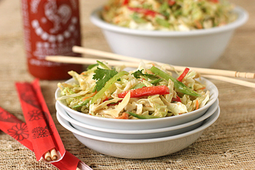 A bowl of fresh cabbage salad with red peppers and green vegetables, garnished with cilantro, sits beside a pair of chopsticks. In the background are a bottle of hot sauce and another bowl of salad.