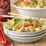 A bowl of fresh cabbage salad with red peppers and green vegetables, garnished with cilantro, sits beside a pair of chopsticks. In the background are a bottle of hot sauce and another bowl of salad.