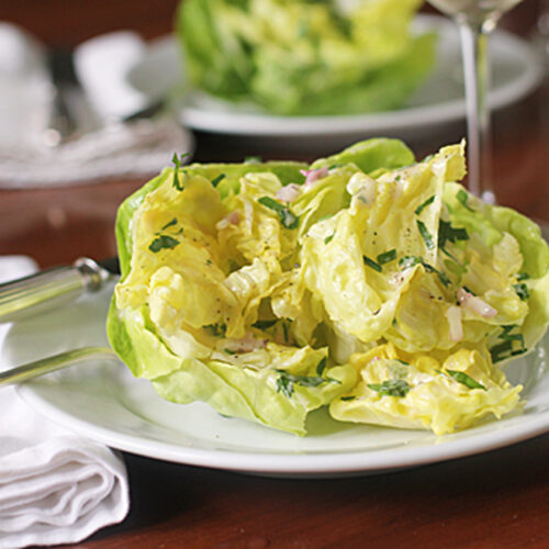 A fresh butter lettuce salad with herbs and sliced onions is served on a white plate with a fork and knife. A folded white napkin and another plate are in the background on a wooden table.