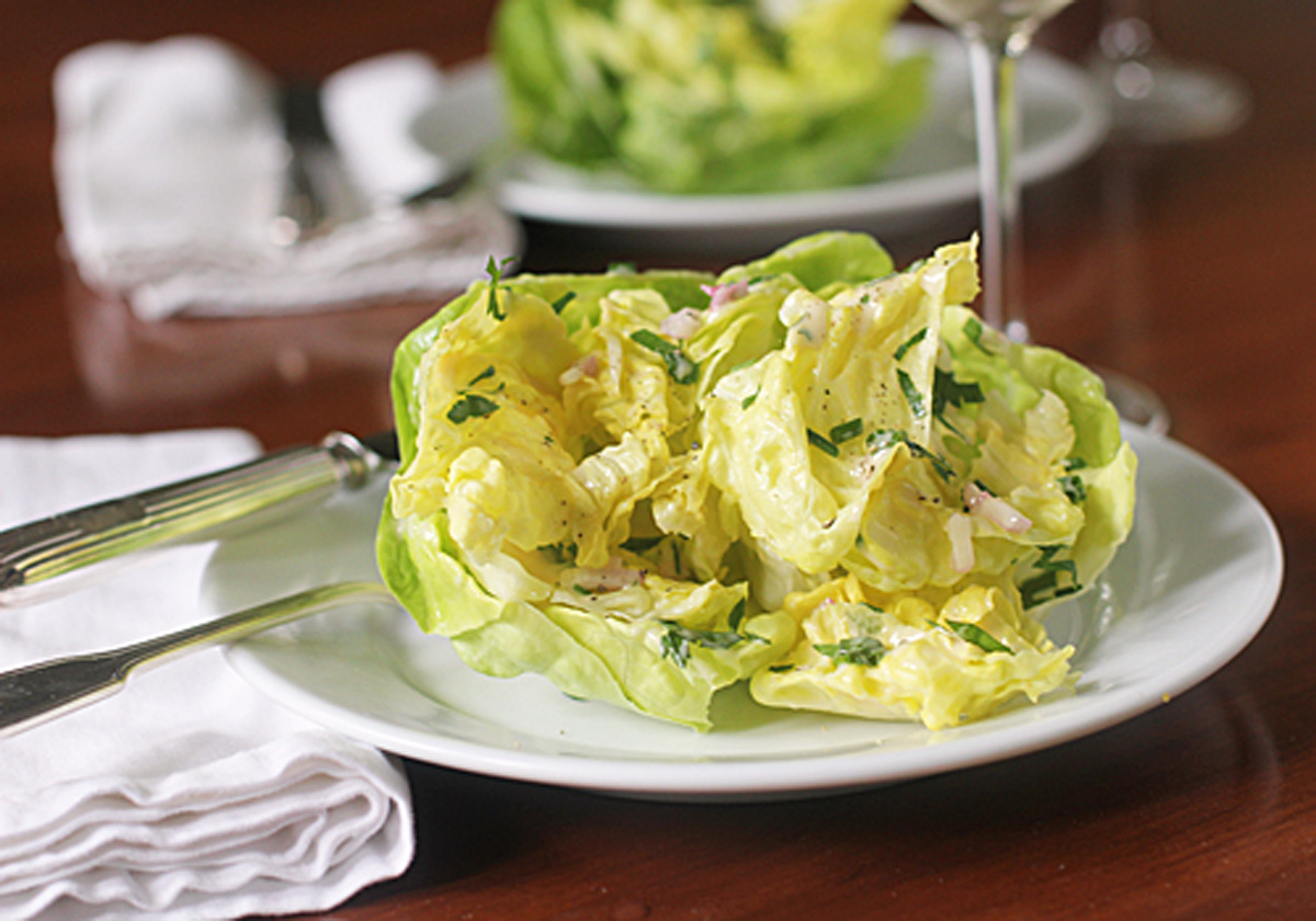 A fresh butter lettuce salad with herbs and sliced onions is served on a white plate with a fork and knife. A folded white napkin and another plate are in the background on a wooden table.