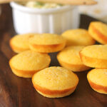 Golden cornbread muffins are arranged on a dark wooden board, with a white ramekin of butter and a wooden spreader in the background.