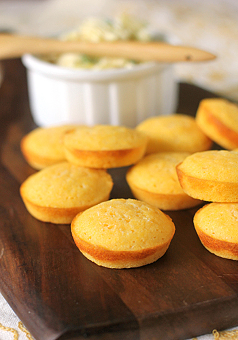 Golden cornbread muffins are arranged on a dark wooden board, with a white ramekin of butter and a wooden spreader in the background.