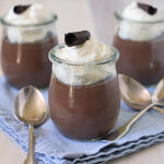 Three glass jars filled with chocolate mousse, topped with whipped cream and a chocolate curl, are placed on a folded blue cloth, surrounded by three metal spoons.