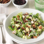 A plate of salad with lettuce, diced chicken, apple pieces, pecans, and dried cranberries, set on a white table next to a fork, a napkin, a green glass of water, and small bowls of pecans and cranberries.
