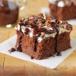 A slice of chocolate cake topped with whipped cream, chopped nuts, and drizzled with chocolate sauce, sits on a piece of parchment paper on a wooden surface. A jar of chocolate sauce is in the background.
