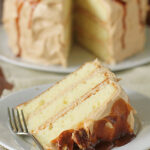 A slice of three-layer vanilla cake with caramel frosting and sauce sits on a plate with a fork. The rest of the cake, similarly frosted and drizzled with caramel, is in the background.