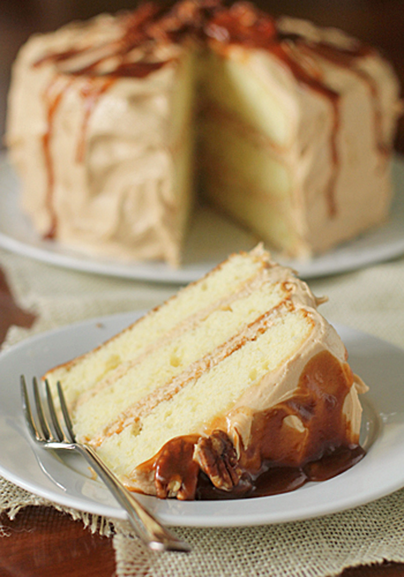 A slice of three-layer vanilla cake with caramel frosting and sauce sits on a plate with a fork. The rest of the cake, similarly frosted and drizzled with caramel, is in the background.
