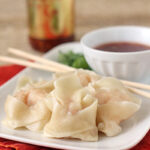 A plate of steamed dumplings with chopsticks rests on an orange napkin, accompanied by a bowl of dipping sauce and a jar of chili sauce in the background.