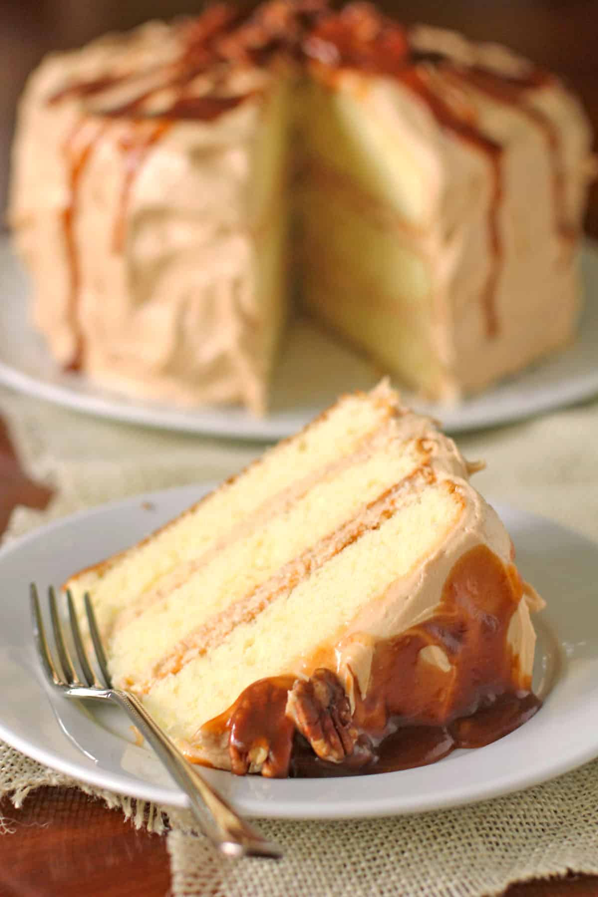 A slice of three-layer cake with caramel frosting and sauce on a white plate, served with a fork. The whole cake is in the background, also topped with caramel.