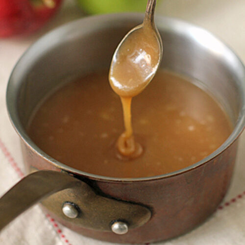 A spoon drizzles caramel sauce above a saucepan filled with caramel, with a red apple and a green apple in the blurred background.