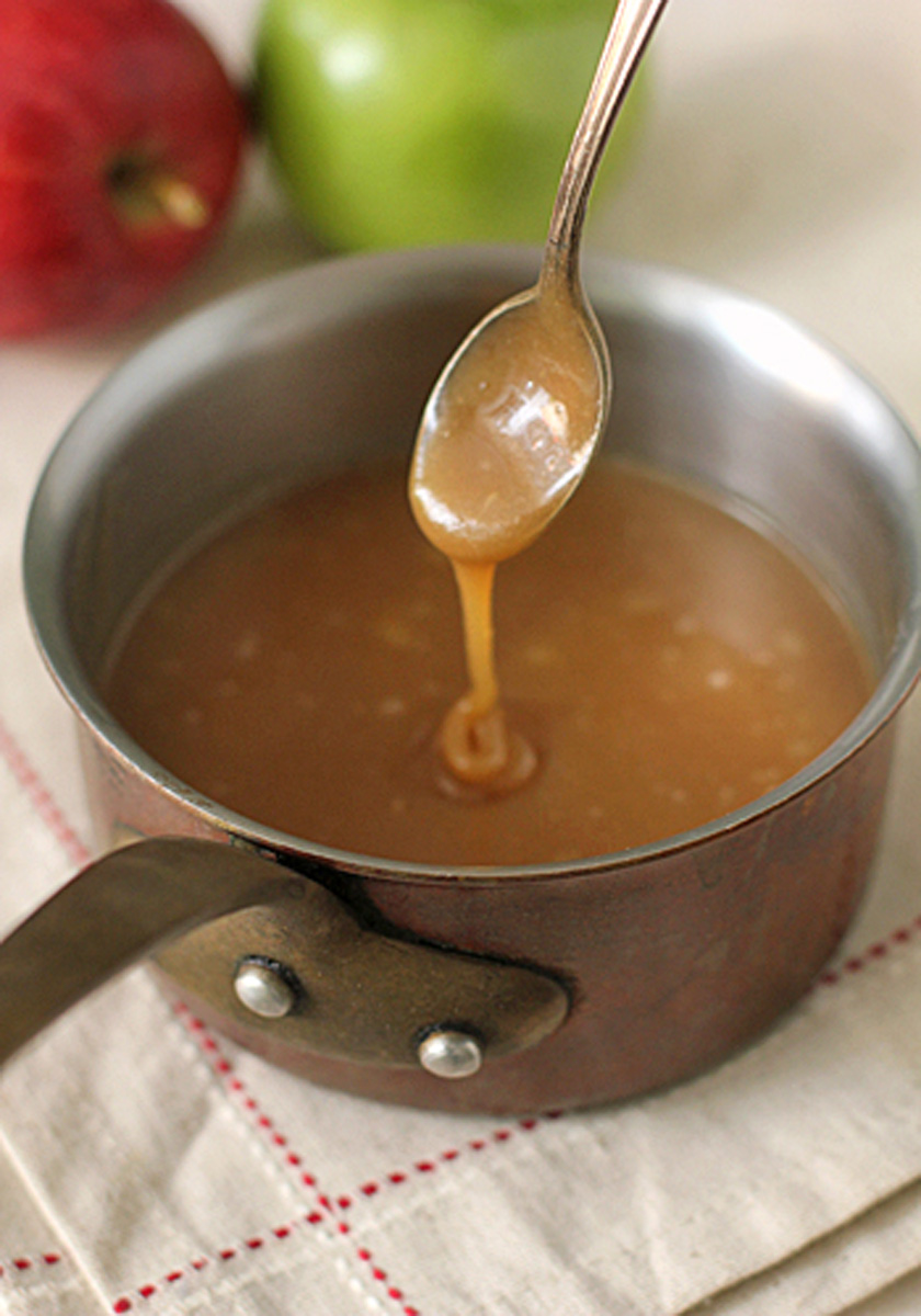 A spoon drizzles caramel sauce above a saucepan filled with caramel, with a red apple and a green apple in the blurred background.