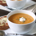 A white bowl of creamy soup topped with a dollop of sour cream and herbs, served on a square white plate with a slice of toasted bread topped with melted cheese and a metal spoon beside it.