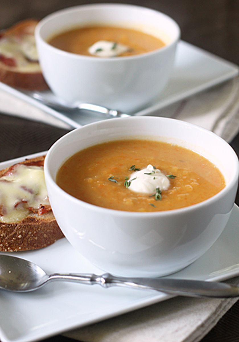 A white bowl of creamy soup topped with a dollop of sour cream and herbs, served on a square white plate with a slice of toasted bread topped with melted cheese and a metal spoon beside it.
