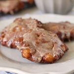 Three glazed apple fritters on a white plate, with a cup of coffee or tea blurred in the background. The fritters have a glossy, sugary coating and sit on a folded blue cloth napkin.