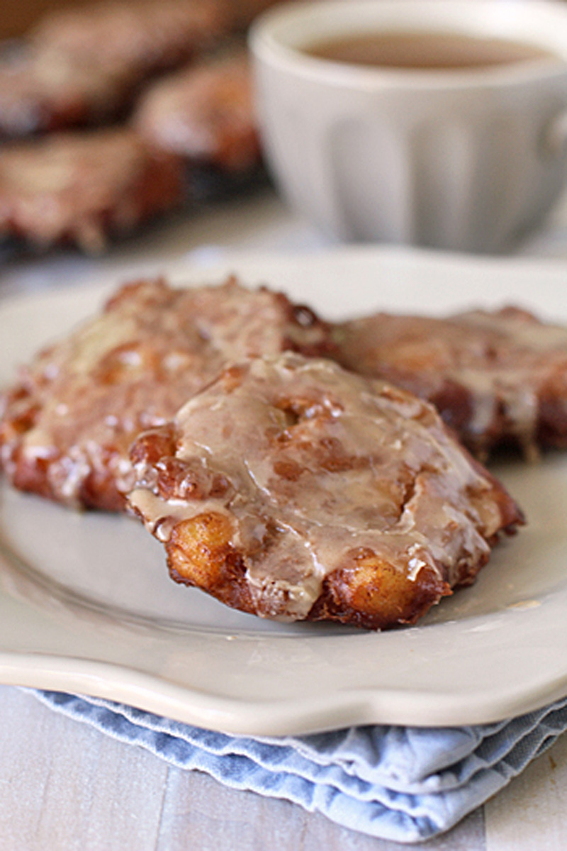 Three glazed apple fritters on a white plate, with a cup of coffee or tea blurred in the background. The fritters have a glossy, sugary coating and sit on a folded blue cloth napkin.