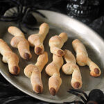 A silver tray holds creepy finger-shaped cookies with almond fingernails, surrounded by fake spiders and a black skull decoration, creating a spooky Halloween scene.