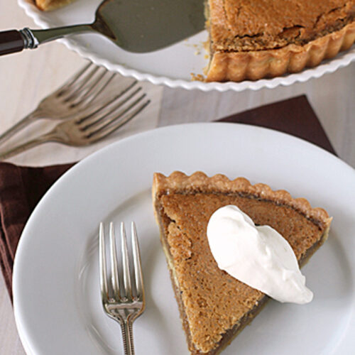 A slice of pie with a dollop of whipped cream on a white plate, next to a fork. The rest of the pie, with one slice missing, is in a dish above, with a pie server and additional forks nearby.