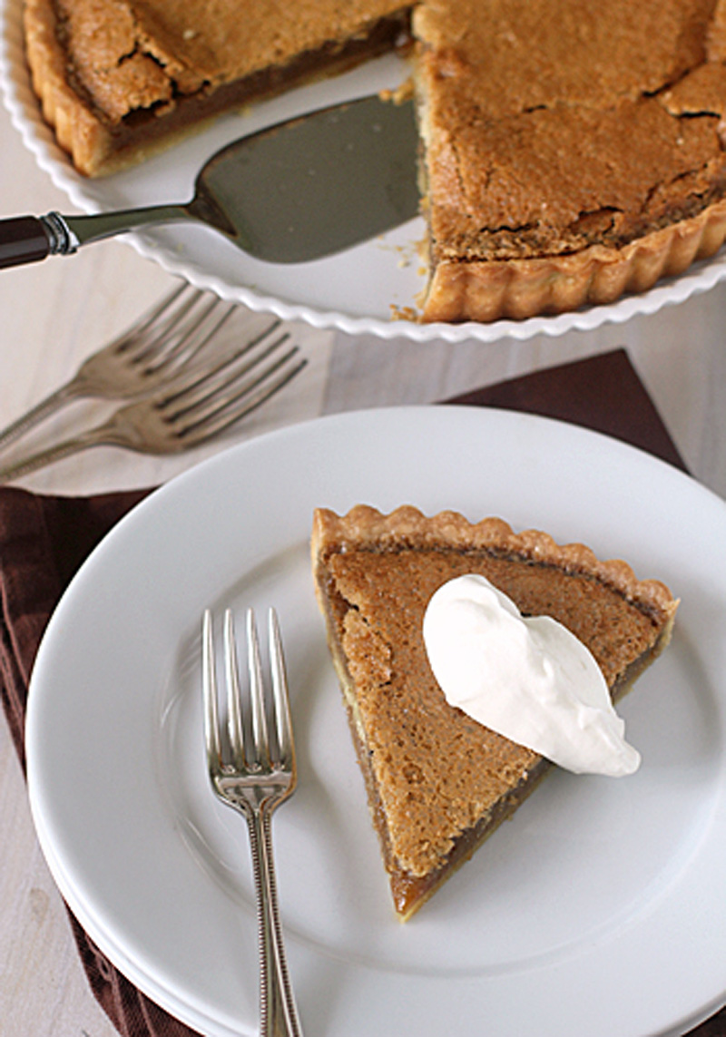 A slice of pie with a dollop of whipped cream on a white plate, next to a fork. The rest of the pie, with one slice missing, is in a dish above, with a pie server and additional forks nearby.
