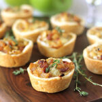 Mini savory tarts filled with caramelized onions and herbs are arranged on a wooden board, with fresh thyme sprigs and green apples in the background.