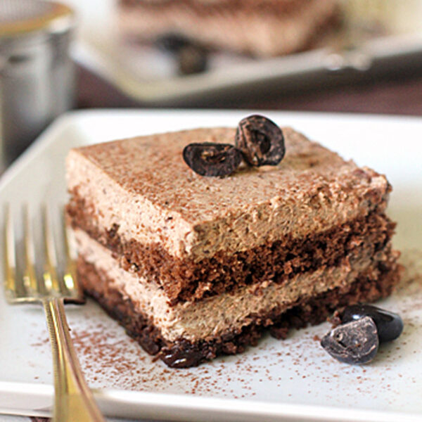 A square slice of layered chocolate mousse cake sits on a white plate with a gold fork, garnished with chocolate pieces and cocoa powder, with another slice blurred in the background.