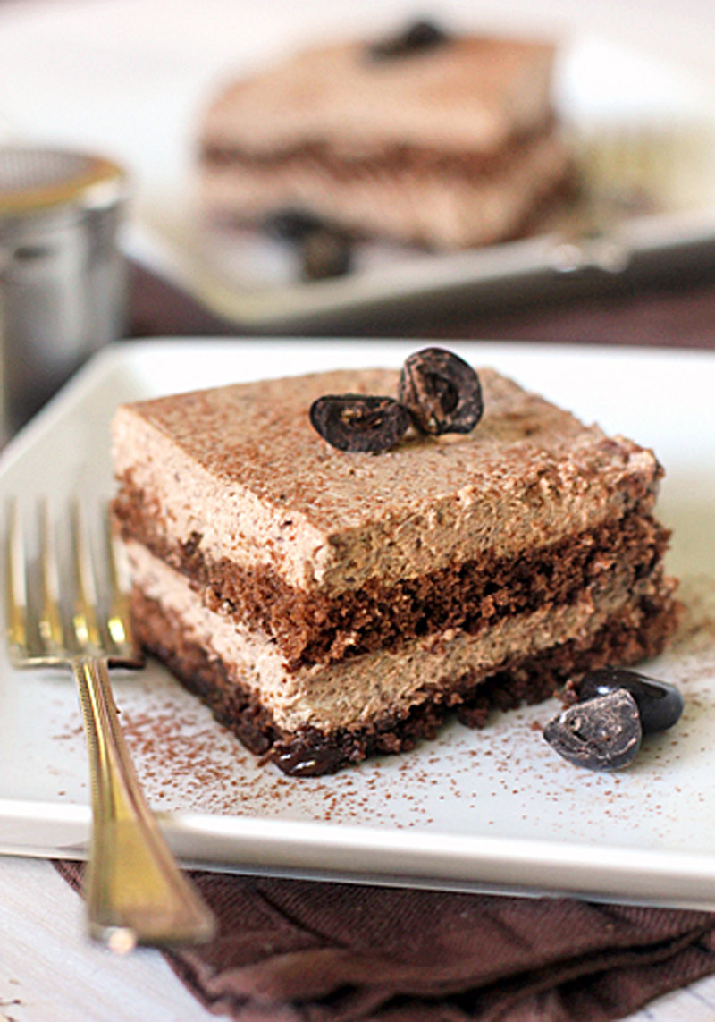 A square slice of layered chocolate mousse cake sits on a white plate with a gold fork, garnished with chocolate pieces and cocoa powder, with another slice blurred in the background.