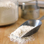 A metal scoop filled with flour rests on a wooden surface, with some flour spilled beside it. A glass jar of flour and a round metal biscuit cutter are in the background.