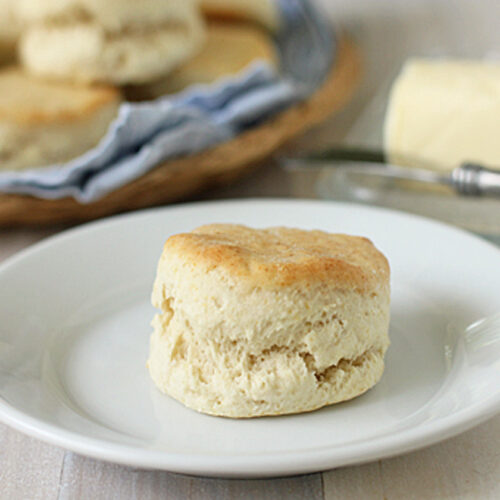 A homemade biscuit sits on a white plate, with a dish of butter and a butter knife in the background. More biscuits are in a basket lined with a blue and white cloth.