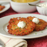 Two crispy potato pancakes topped with sour cream and chopped chives on a white plate, with bowls of applesauce and more sour cream in the background.
