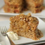 A slice of moist cake topped with chopped nuts and caramel glaze sits on a white plate with a dollop of cream and a fork; the rest of the cake is in the background.