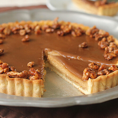 A caramel tart with a slice removed, topped with candied nuts and set on a round metal plate. The tart has a smooth caramel layer and a golden crust, with a slice served on a separate plate in the background.