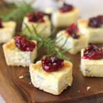 Small square bites of blue cheese tart topped with red cranberry sauce are arranged on a wooden serving board, garnished with sprigs of fresh rosemary in the background.