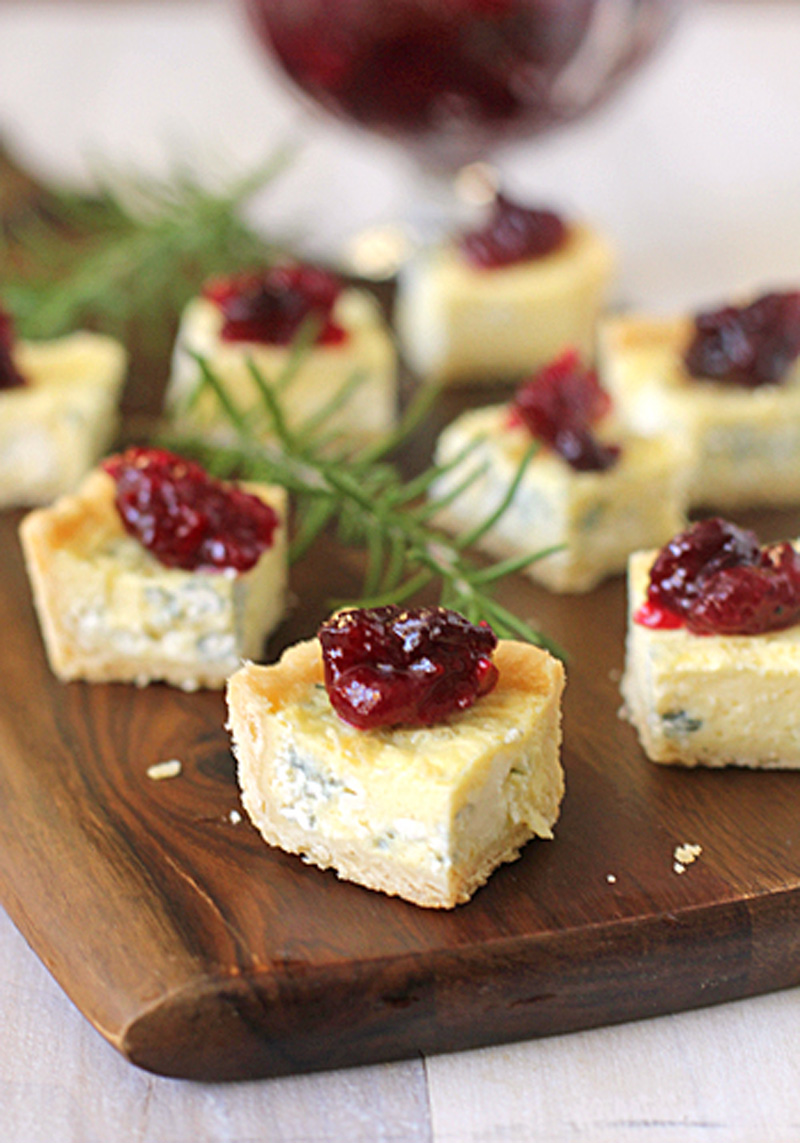 Small square bites of blue cheese tart topped with red cranberry sauce are arranged on a wooden serving board, garnished with sprigs of fresh rosemary in the background.