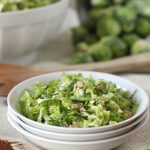 A white bowl filled with shredded Brussels sprouts salad, topped with nuts, sits on stacked white plates. In the background, a colander with whole Brussels sprouts is visible.
