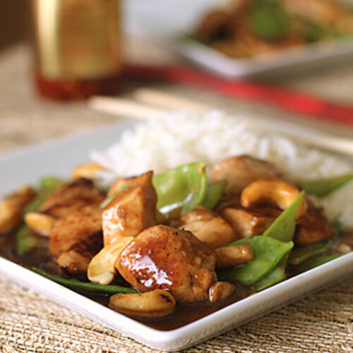 A square white plate holds a serving of rice, stir-fried chicken, snow peas, and cashews in sauce. Wooden chopsticks rest nearby, and a jar of sauce is blurred in the background on a woven mat.
