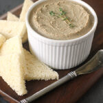 A white ramekin filled with smooth pâté, garnished with a small sprig of thyme, is placed on a wooden board beside several triangular slices of white bread and a silver butter knife.