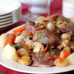 A plate of coq au vin with chicken, mushrooms, pearl onions, carrots, and bacon, garnished with parsley. Wine glasses, a wine bottle, plates, and a gravy boat are in the blurred background.