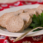 Slices of pale sausage-like meat, possibly a type of luncheon meat or pate, arranged on a white plate with a sprig of parsley and a fork, set on a red patterned cloth.