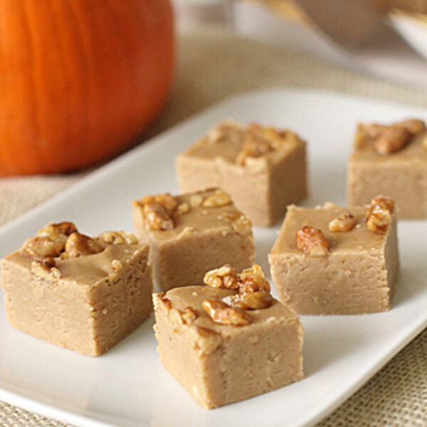 Squares of light brown fudge topped with chopped nuts are arranged on a white rectangular plate. A pumpkin sits in the background on a beige tablecloth, suggesting an autumn or pumpkin-flavored dessert.