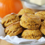 A plate of golden, fluffy pumpkin biscuits sits on a cloth napkin. In the background, a whole pumpkin and a dish of butter are visible on a wooden table.