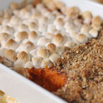 A casserole dish with sweet potato casserole topped half with toasted marshmallows and half with a crumbly pecan streusel, with a scoop missing to reveal the orange sweet potato layer.