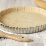 A fluted tart pan with raw pastry dough pressed into it sits on a lightly floured surface. A wooden rolling pin and a pastry brush are nearby.