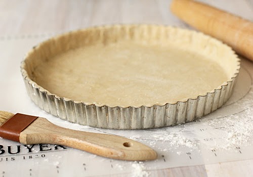 A metal tart pan filled with unbaked pie crust sits on a floured surface, with a rolling pin and a pastry brush nearby.
