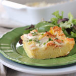 A slice of cheesy breakfast casserole garnished with chives is served on a green plate with a side of mixed greens. A fork and knife rest on a napkin beside the plate.