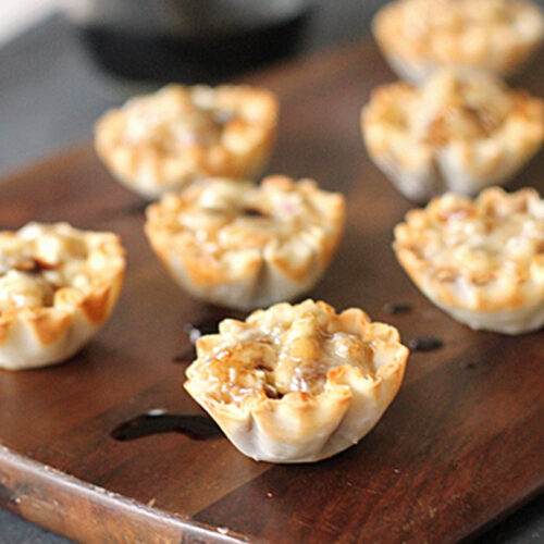 Mini phyllo pastry cups filled with a baked mixture, possibly cheese and nuts, are arranged on a wooden board with a drizzle of sauce and a glass container in the blurred background.