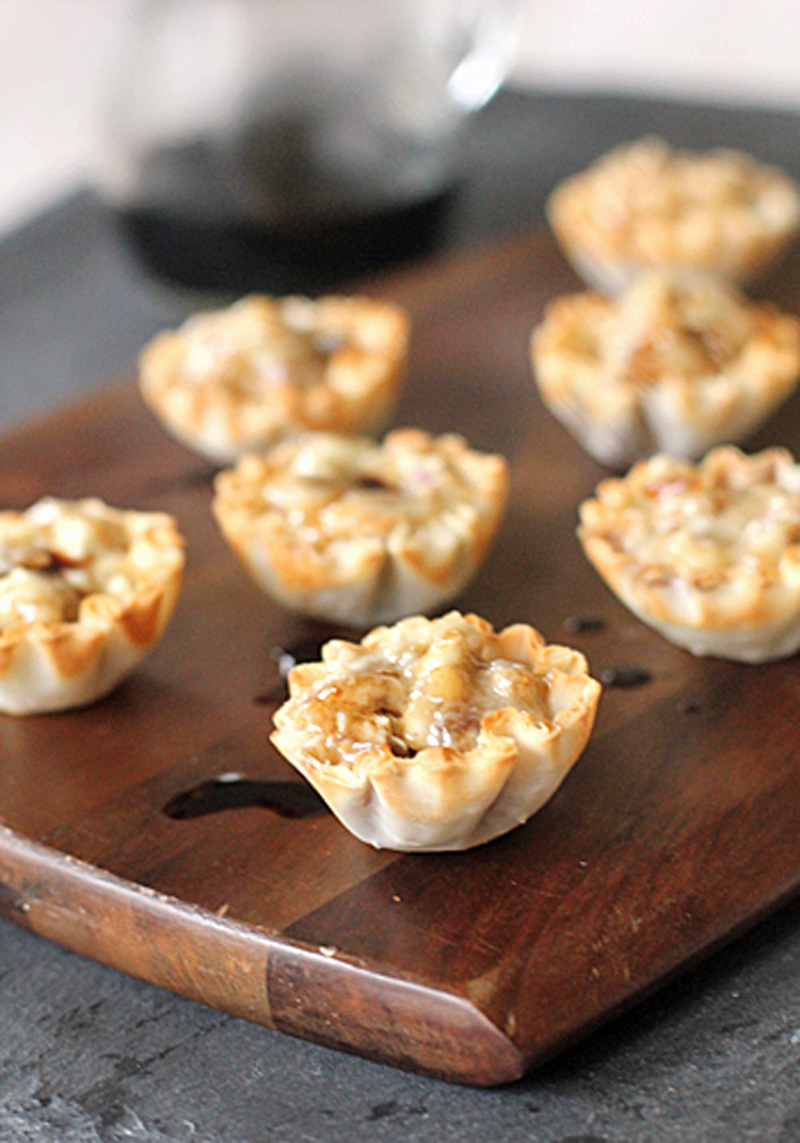 Mini phyllo pastry cups filled with a baked mixture, possibly cheese and nuts, are arranged on a wooden board with a drizzle of sauce and a glass container in the blurred background.