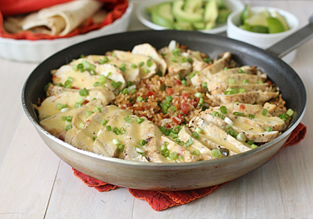 A skillet filled with sliced grilled chicken, chopped green onions, and a cheesy tomato rice mixture, served on a red cloth. In the background, tortillas, sliced avocado, and lime wedges are visible.