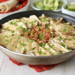 A skillet filled with sliced grilled chicken, chopped green onions, and a cheesy tomato rice mixture, served on a red cloth. In the background, tortillas, sliced avocado, and lime wedges are visible.
