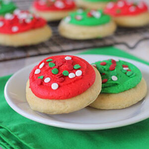 Two frosted sugar cookies with red and green icing and festive sprinkles sit on a white plate, with more decorated cookies on a wire rack in the background. A green napkin is under the plate.