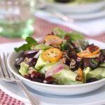A fresh salad with mixed greens, red onion, dried cranberries, candied pecans, and orange slices is served in a white bowl on a plate, with a fork beside it on a red checkered tablecloth.